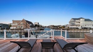 A waterfront deck with wooden flooring and black lounge chairs, overlooking a marina with boats and houses. - Waterfront Mantoloking Home with Private Dock w/Pool very close to Ocean Beach  (Mantoloking)