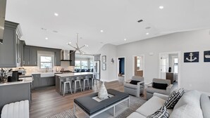 A modern kitchen with grey cabinets, a central island, and bar stools. There is a dining area with a white table and chairs. The living room features a grey sofa, a coffee table, and a patterned rug. - Waterfront Mantoloking Home with Private Dock w/Pool very close to Ocean Beach  (Mantoloking)