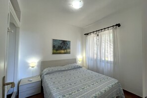 Ein Schlafzimmer mit einem Bett, einem Nachttisch, einer Wandleuchte, einem Gemälde und einem Fenster mit Vorhängen.