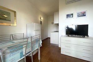 Una cocina compacta con refrigerador, una mesa de comedor con sillas, un televisor sobre un mueble bajo y un mueble auxiliar pequeño.