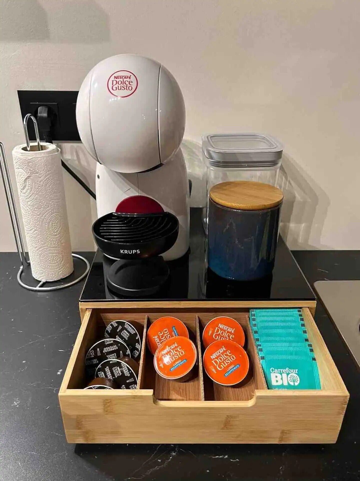 Una cafetera, un dispensador de toallas de papel, un recipiente y un cajón con cápsulas de café.
