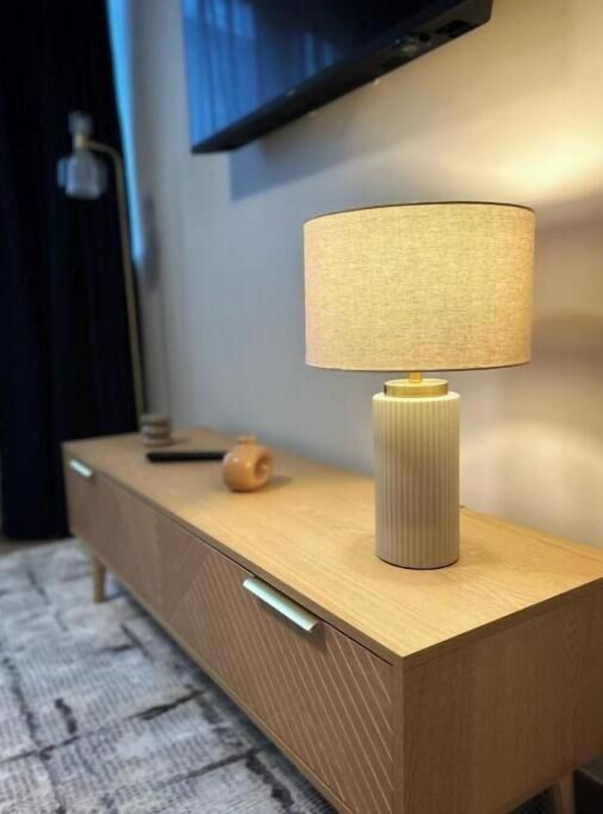 En modern byrå i trä med en lampa med intressant yta ovanpå, en liten vas och en fjärrkontroll.