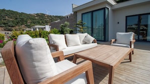 Une terrasse en bois avec un canapé et des fauteuils, une table et une grande porte vitrée donnant sur un bâtiment moderne.