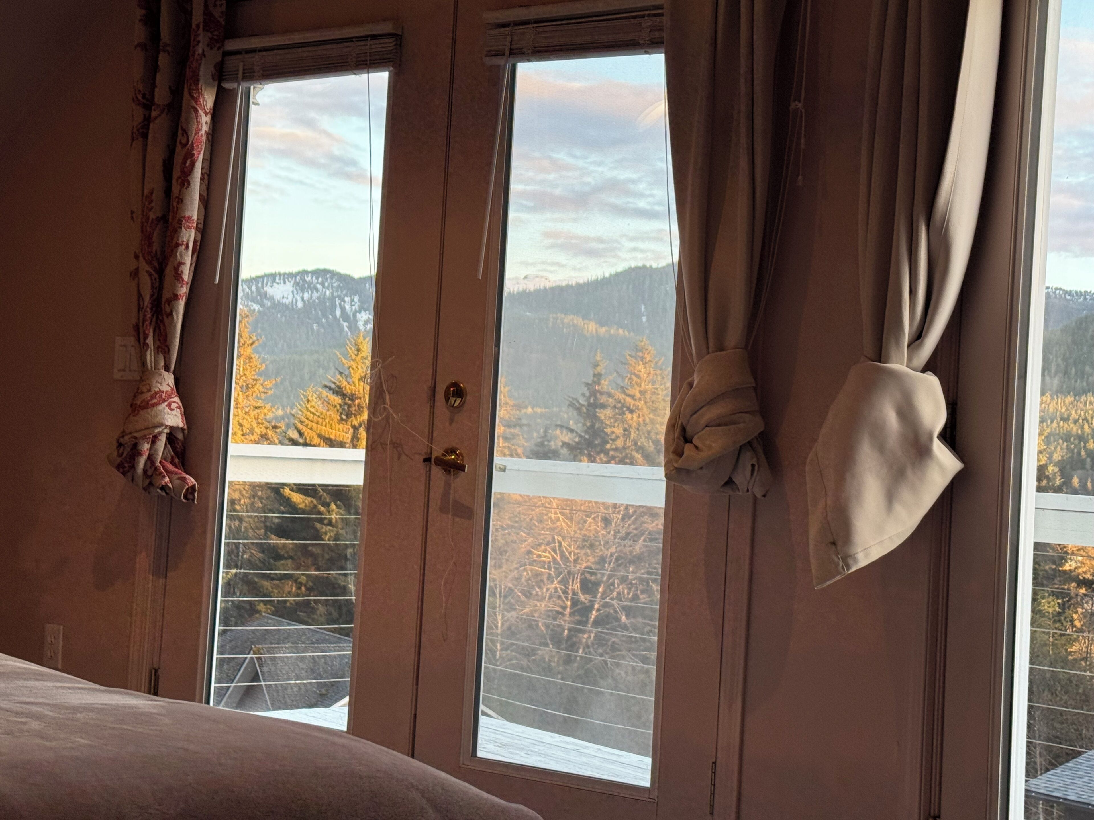 A room with a large window, curtains, and a view of trees and mountains.