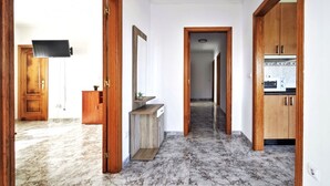Un couloir avec des portes en bois, un sol en marbre, un téléviseur fixé au mur et un coin cuisine visible à travers une porte ouverte.