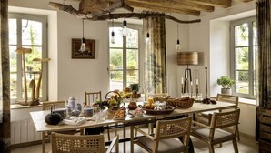 Comedor rústico con vigas de madera, ventanas amplias y una variedad de vajilla y alimentos.