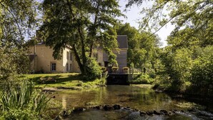 Una escena tranquila a orillas del río, con un puente de piedra tradicional, abundante vegetación y un edificio histórico de fachada de piedra y múltiples ventanas.