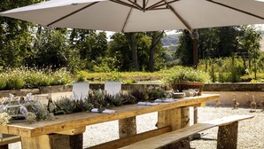 Un área de comedor al aire libre con una mesa de madera dispuesta para una comida, rodeada de follaje y un paraguas grande que proporciona sombra.