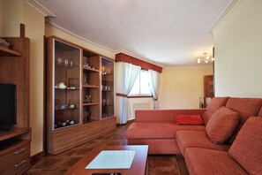 Ein Wohnzimmer mit einer Couch, einem Holzschrank mit Glastüren, einem Fernseher und einem Fenster mit Vorhängen.