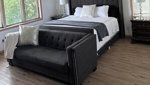 Een slaapkamer met een groot bed, een donkere gestoffeerde bank, een plafondventilator en een raam met jaloezieën.