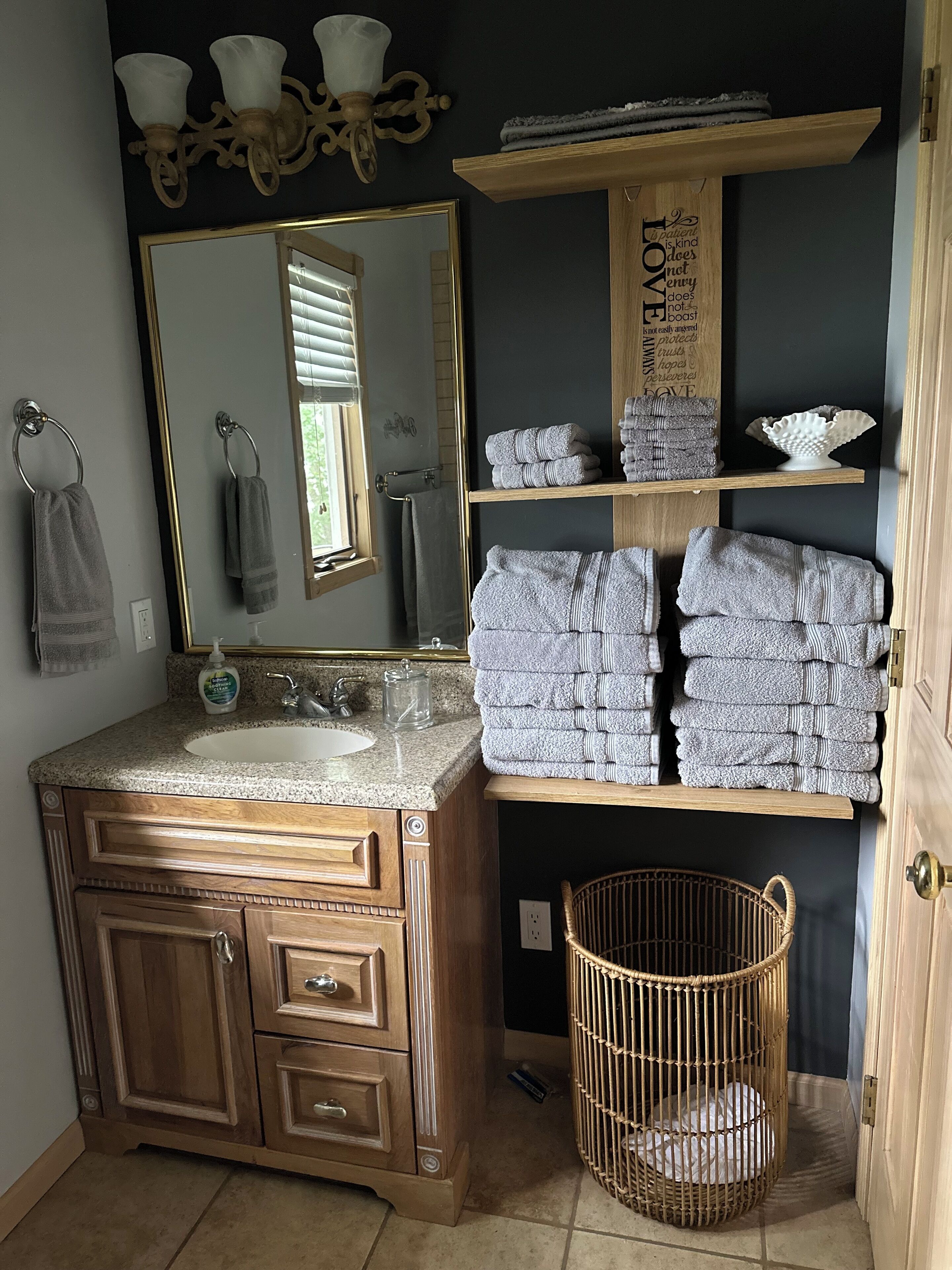 Une salle de bain avec un meuble-lavabo en bois, un grand miroir et un porte-serviettes. On y trouve un panier en osier et une étagère avec des serviettes pliées.