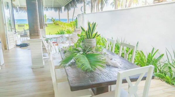 A dining area with a table set for two, surrounded by white chairs, a potted plant, and a view of the ocean.