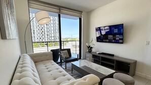 A modern living room with a sofa, a coffee table, a TV, and a balcony with a view of a building. - Walk to Downtown Doral / Resort Pool Free Parking (Doral)