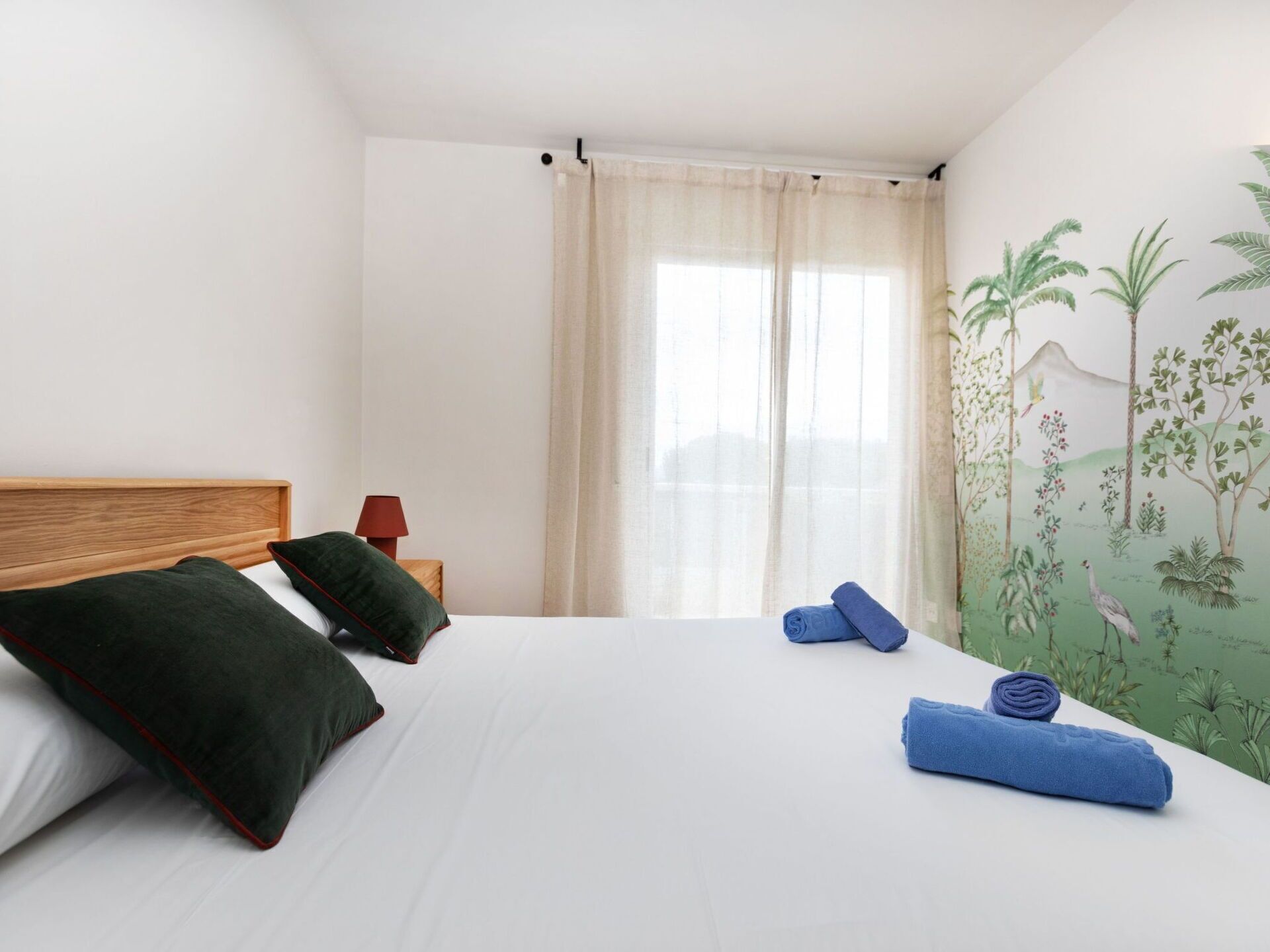 Ein Schlafzimmer mit Bett, Kissen, einer Lampe und einer Wand mit einem Wandgemälde zum Thema Natur.