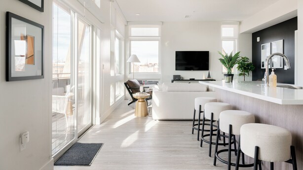 A modern kitchen with a large island, bar stools, a flat-screen TV, and a view of the city through large windows. - Skyline Panorama | 2BR LoHi Loft | Dog-Friendly (Denver)