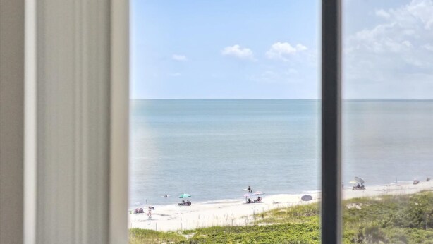 A beach view through a window, including the ocean, beach, and some people. - Charming Oceanfront Condo with Breathtaking Views, Shuffleboard & Heated Pool! 🌊 (Cape Canaveral)