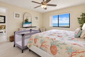 Een slaapkamer met een bed, een plafondventilator, een televisie, een raam met uitzicht op de oceaan en een wieg.