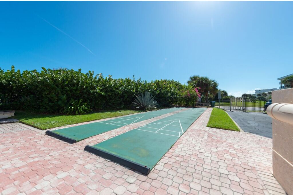 A sunny outdoor area with a paved walkway, green exercise mats, and a hedge.