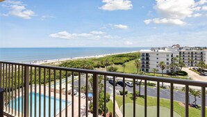 A view from a balcony overlooking a beach, a swimming pool, and a parking area with a multi-story building in the background. - Charming Oceanfront Condo with Breathtaking Views, Shuffleboard & Heated Pool! 🌊 (Cape Canaveral)