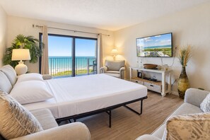 A hotel room with a bed, a TV, a balcony with a view of the beach, and a dining area with a table and chairs. - Spacious 3BR Corner Condo | Perfect for Groups + Ocean Views from Every Angle! 🌴 (Cape Canaveral)