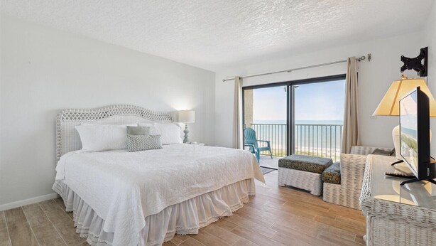 A bedroom with a large bed, a balcony with a view, and a TV on a side table. - Spacious 3BR Corner Condo | Perfect for Groups + Ocean Views from Every Angle! 🌴 (Cape Canaveral)