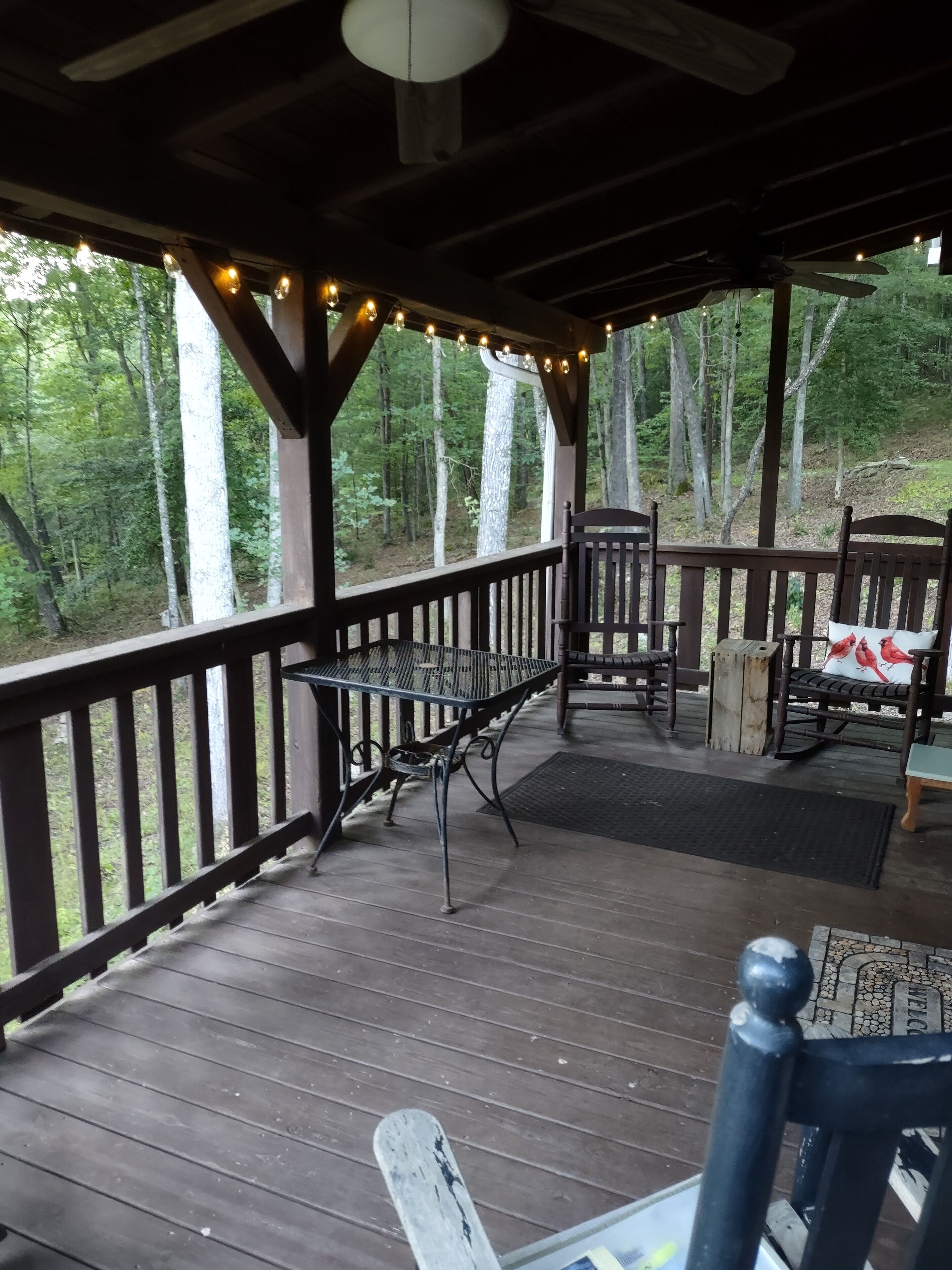 A wooden deck with a table and chairs, string lights, and a view of a forest.