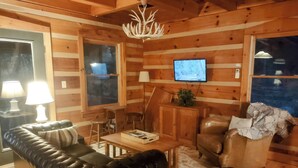 L’intérieur d’un chalet confortable, avec des murs en rondins de bois, un canapé en cuir, une table basse en bois, un téléviseur et une cheminée.
