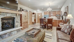 A spacious living area with a stone fireplace, a dining table with chairs, a sofa, and a kitchen in the background. - 1 Bedroom Condo (Keystone)