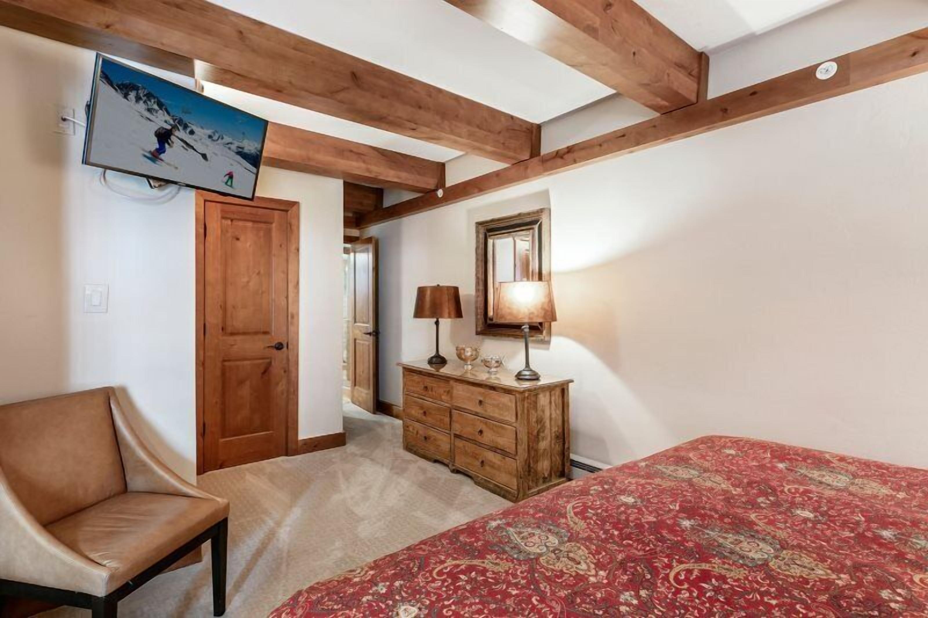 Un dormitorio con techo de madera, una cama con colcha estampada roja, un aparador de madera, una silla y un televisor montado en la pared.