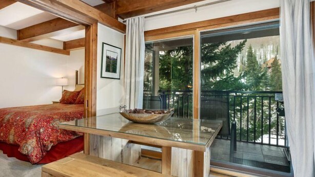 Ein Schlafzimmer mit Glastisch, roter Bettdecke und einem Balkon mit Blick auf Bäume.