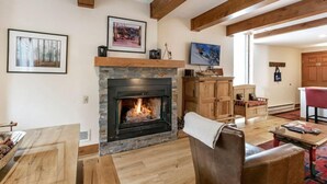 Ein gemütliches Wohnzimmer mit Kamin, Holzbalken, einem Steinumbau für den Kamin, einer Holzvitrine, einem Fernseher und einem roten Sessel.