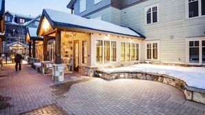 En snötäckt byggnad med en fasad av sten och stora fönster, omgiven av ett plattbelagt område och andra byggnader.