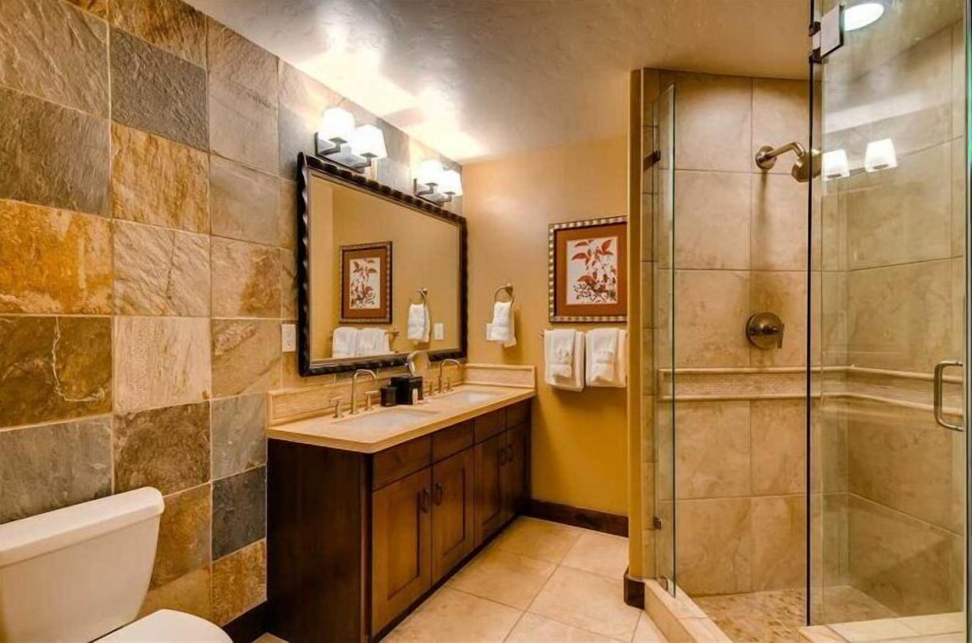 A bathroom with a stone tile wall, a large mirror, a glass-enclosed shower, and a wooden vanity.