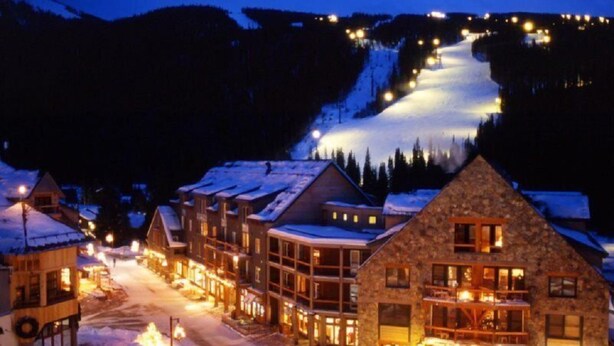 Un pueblo cubierto de nieve al caer la noche, con edificios iluminados y una pista de esquí iluminada.