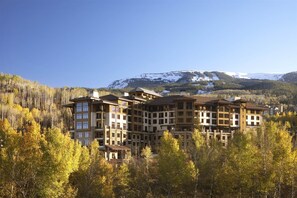 Un hôtel à plusieurs étages, doté d’une façade en pierre, de grandes fenêtres et de balcons, entouré d’un feuillage automnal et surplombant un paysage montagneux.