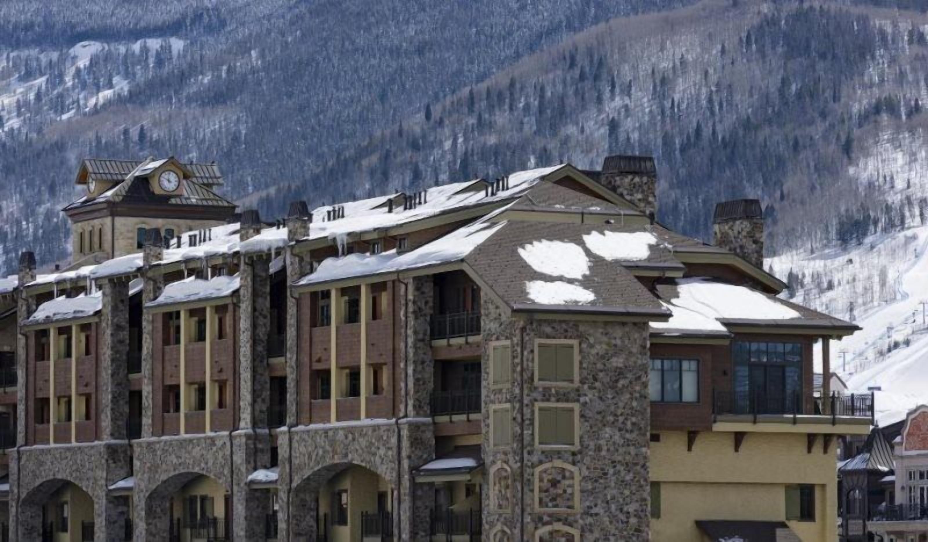 A multi-story hotel with stone and wood architecture, featuring balconies and large windows, set against a mountainous backdrop.