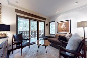 Uma sala de estar moderna com um sofá, duas poltronas, uma mesa de centro e uma janela grande com vista para árvores.
