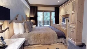 A bedroom with a large bed, a wooden wardrobe, and a balcony with a view of trees.