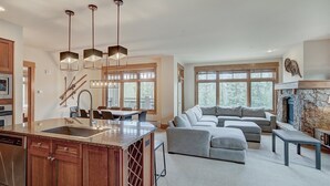 Uma sala de estar espaçosa com um amplo sofá modular cinza, uma cozinha com armários de madeira e bancadas de granito, e uma lareira de pedra.