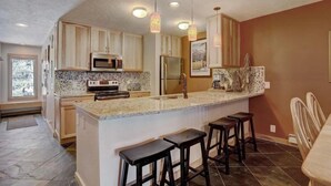 Una cocina con encimeras de granito, armarios de madera, una isla central con taburetes e iluminación colgante.