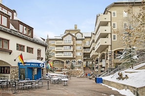 A snow-covered outdoor area with tables and chairs, a Blue Moose Pizza restaurant, and a building with multiple balconies. - 3 Bedroom - St James (Avon)