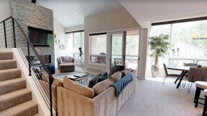 Una sala de estar moderna con chimenea de piedra, una escalera, un sofá y un comedor con vistas al exterior.