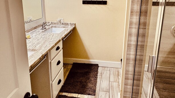 A bathroom with a marble countertop, a framed picture on the wall, a black rug, and a shower with glass doors. - Beach Manor 0309 (Miramar Beach)