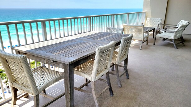 Varanda com mesa de madeira e cadeiras de vime, com vista para uma praia e água turquesa.