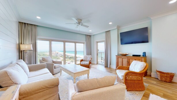 A spacious living room with a large window offering a view of the beach, a ceiling fan, and a flat-screen TV mounted on the wall. - Beach Manor 1005 (Miramar Beach)