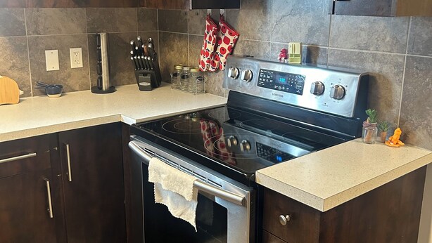 A kitchen with dark wooden cabinets, a stainless steel stove, and a tiled backsplash. - Basement suite with full kitchen  (Airdrie)