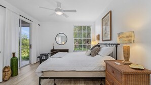 Een slaapkamer met een groot bed, een plafondventilator, een raam met uitzicht, een nachtkastje met een lamp en een spiegel aan de muur.