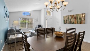 A dining area with a wooden table, chairs, and a chandelier. There is a window with blinds, a sofa, and a lamp.