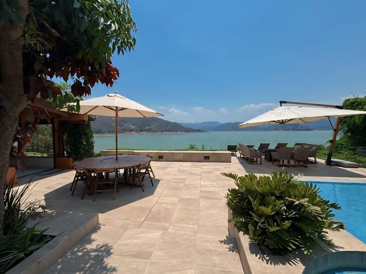 Un área junto a la piscina con mesa y sillas, sombrillas y vistas a un lago.