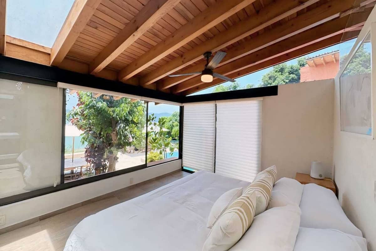 A bedroom with a large bed, wooden ceiling beams, a ceiling fan, and a view of a swimming pool and trees outside.